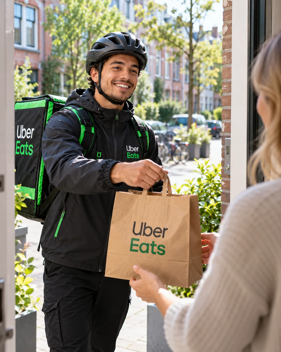 Uber Eats-bezorger in zwarte jas met helm overhandigt een bestelling in een papieren tas aan een klant bij de voordeur
